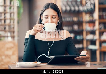Beautiful, happy and relaxed student with tablet drinking coffee, listening to a podcast and music on earphones in a cafe. Woman online and watching Stock Photo