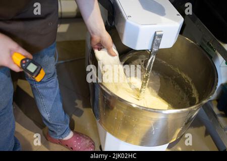 The baker kneads bread in a dough mixer. The process of making bread in an artisan bakery. Measuring the temperature of the dough. Front view. Stock Photo
