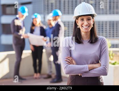 Were confident for a reason. a young businesswoman standing with her arms folded and wearing a hardhat while her colleagues stand behind her. Stock Photo