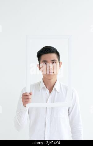 Young unsmiling Asian man looking at camera through picture frame Stock Photo