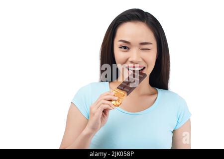 Waist-up portrait of cheerful Asian woman giving wink while standing against white background and biting chocolate bar Stock Photo