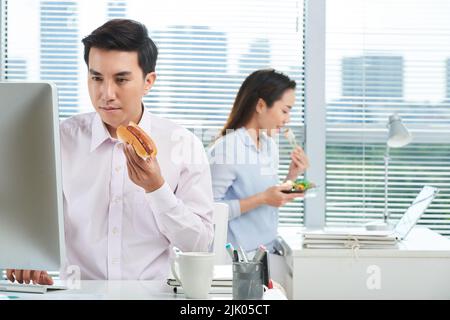 Busy open plan office: concentrated Asian manager wearing white shirt working on computer and eating hot dog while his pretty colleague sitting at workplace and enjoying sushi Stock Photo