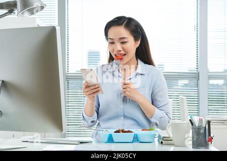 Attractive Asian entrepreneur eating delicious salad while sitting at office desk and texting with friend on smartphone, panoramic window on background Stock Photo