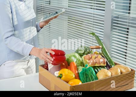 Online grocery delivery: unrecognizable young woman using digital tablet while checking amount of products in her order, cardboard box standing on table Stock Photo
