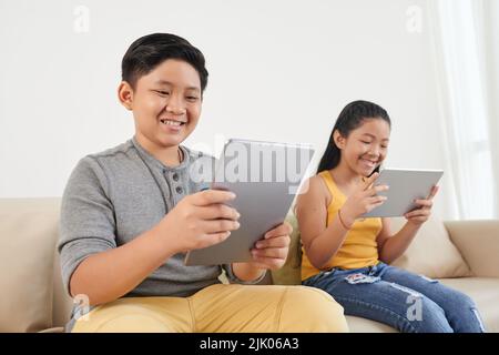 Vietnamese brother and sister sitting on sofa and watching movies on tablet computers Stock Photo