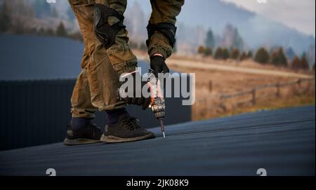 Male builder installing black corrugated iron sheet used as facade of future cottage. Man worker building wooden frame house. Carpentry and construction concept. Stock Photo
