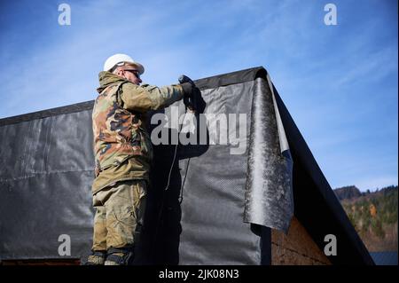 Male builder installing waterproof membrane on the wall of future cottage. Man worker building wooden frame house with blue sky on background. Carpentry and construction concept. Stock Photo