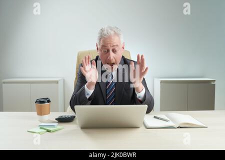 Shocked mature businessman wearing classical suit looking on laptop screen and shouting, interior of modern office on background Stock Photo