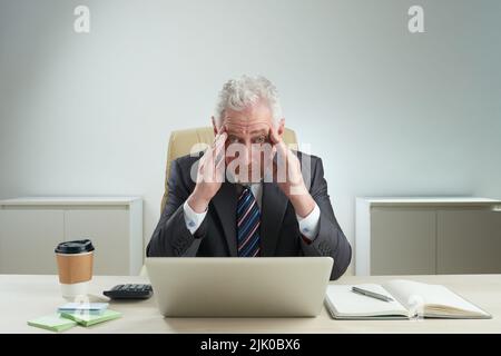 Tired senior white collar worker looking at camera while rubbing his temples and sitting at office desk, waist-up portrait shot Stock Photo