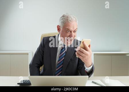 Waist-up portrait of furious senior entrepreneur wearing classical suit sitting at office desk and shouting while holding ringing phone in hand, office interior on background Stock Photo