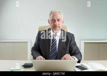 Confident elderly white collar worker looking at camera while sitting at office desk and analyzing statistic data with help of laptop, waist-up portrait shot Stock Photo