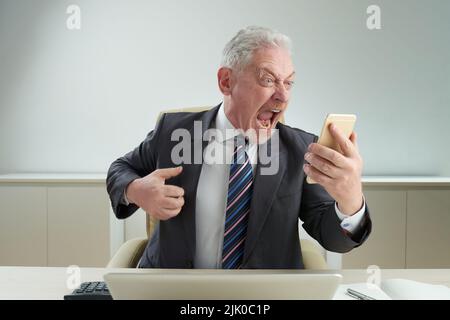 Angry senior white collar worker wearing classical suit sitting at office desk and shouting at smartphone after stressful talk with his boss, office interior on background Stock Photo