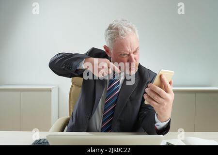 Furious elderly businessman wearing classical suit shaking finger and shouting at ringing smartphone while sitting at office desk, waist-up portrait shot Stock Photo
