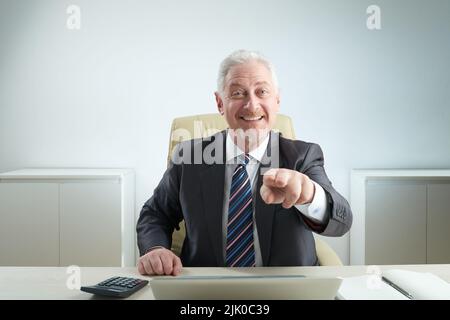 Cheerful aged businessman wearing classical suit pointing at camera while distracted from work in spacious office, waist-up portrait shot Stock Photo