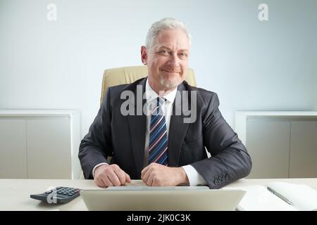 Bearded elderly entrepreneur in formalwear sitting at workplace and looking at camera with wide smile, office interior on background Stock Photo