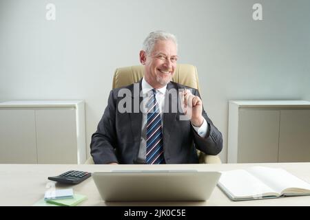 Portrait shot of cheerful senior businessman wearing classical suit sitting at office desk and looking at camera with wide smile Stock Photo