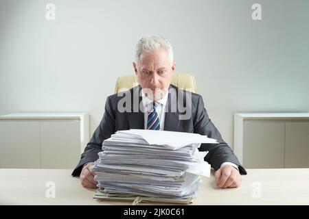 Shocked senior manager wearing classical suit looking at pile of documents while sitting at office desk, waist-up portrait shot Stock Photo