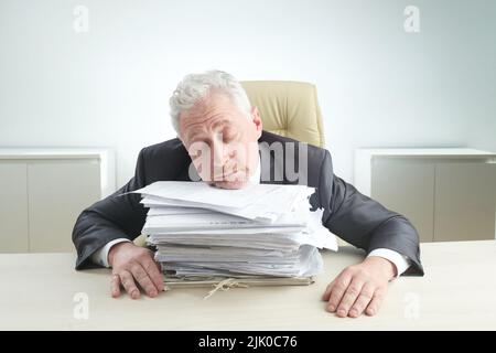 Exhausted elderly manager resting head on pile of documents while sitting at office desk overwhelmed by work, portrait shot Stock Photo
