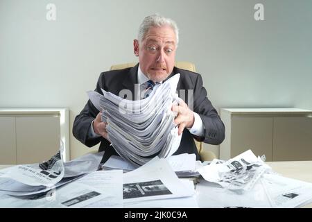 Surprised aged white collar worker wearing classical suit holding pile of documents in hands while sitting at office desk covered with crumpled paper Stock Photo