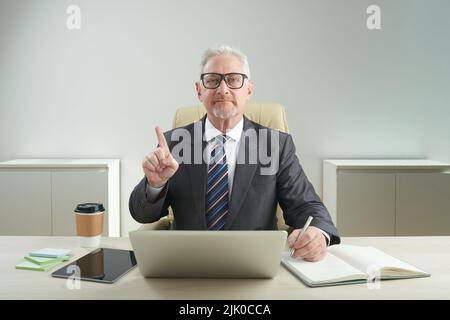Waist-up portrait of serious aged entrepreneur wearing eyeglasses looking at camera and shaking finger while sitting at office desk and working on important project Stock Photo