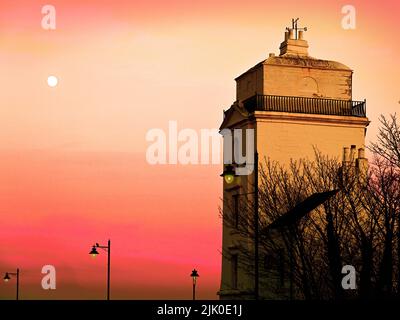 North Shields High Light Lighthouse 1808 at moonrise with golden and scarlet sky and lit street lamps Stock Photo