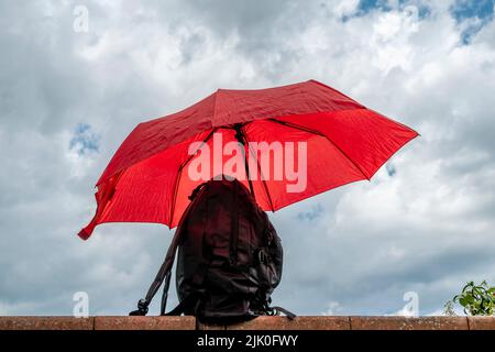 A black backpack protected by a red umbrella against a cloudy sky that threatens rain Stock Photo