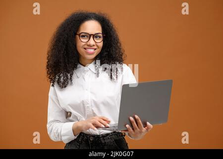 Smart and confident African-American woman in smart casual wear using laptop isolated on brown background, female office employee or student typing, web browsing Stock Photo