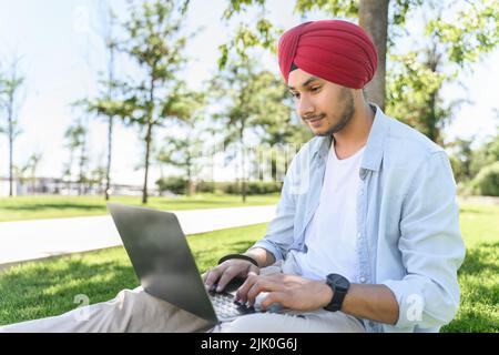 Male Indian student wearing red traditional national turban using laptop computer sitting on the lawn, preparing homework near campus outdoors, hindu guy with typing, replying emails Stock Photo