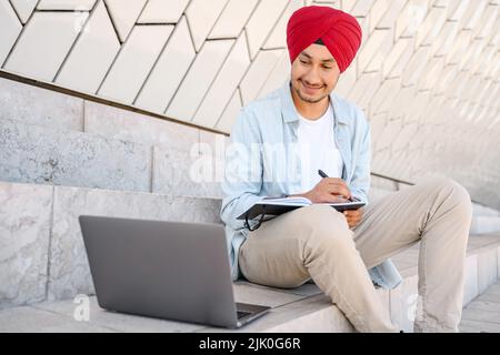 Indian student man in traditional turban sitting outdoors and using laptop for learning on the distance, hindu guy watching webinar and taking notes, male student studying online, e-learning Stock Photo