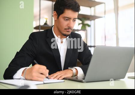 Serious intelligent concentrated young businessman, ceo, manager working on laptop, doing online market research, working on project, creating plan or checking financial statement, taking notes Stock Photo