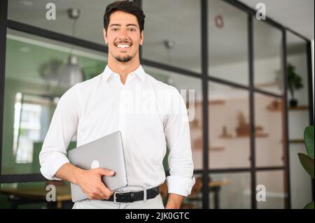 Waist up portrait of the proud businessman carrying laptop in hand and smiling, Latin freelancer man posing in modern loft office with glass partition on the background, looks at camera Stock Photo