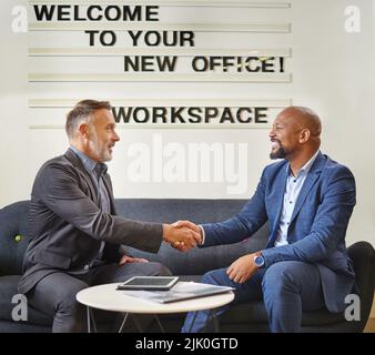 New office, new employees. two businessmen shaking hands while sitting together in a modern office. Stock Photo