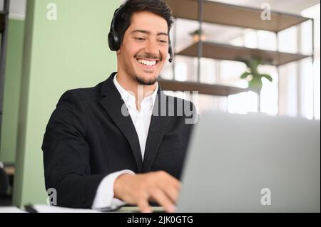 Portrait of latin millennial young businessman wearing headset looking on the laptop, customer service representative sitting in modern open space office, talking online on the distance Stock Photo