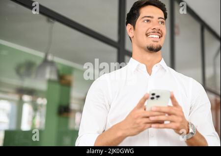 Smiling hispanic business man with a smartphone in hand looks away standing indoor, male office employee, entrepreneur using smartphone, chatting and texting online Stock Photo