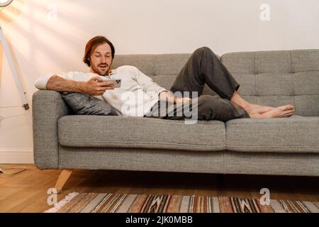 Ginger bristle man using mobile phone while resting on couch at home Stock Photo