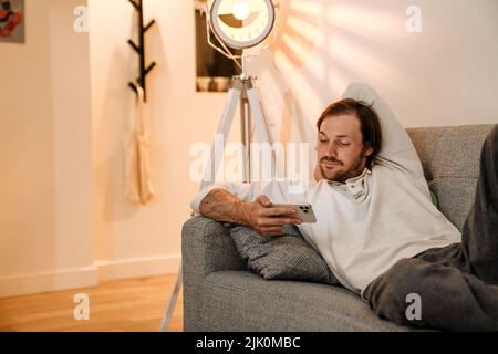 Ginger bristle man using mobile phone while resting on couch at home Stock Photo