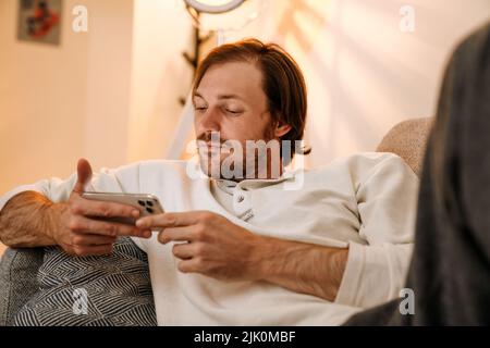 Ginger bristle man using mobile phone while resting on couch at home Stock Photo