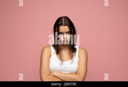 Beautiful girl with brunette long hair frowning her face in displeasure, wearing white t-shirt, keeping arms folded. Attractive young woman in closed Stock Photo