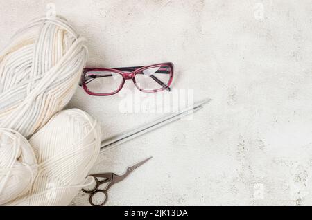 Light beige knitting wool, knitting needles and eyeglesses on conkrete background. Female hobby knitting. Soft woolen yarns for knitting warm clothes Stock Photo