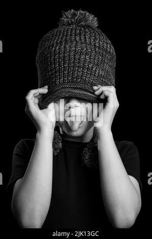 A grayscale vertical shot of a little boy hiding his eyes under a hat and showing a tongue Stock Photo