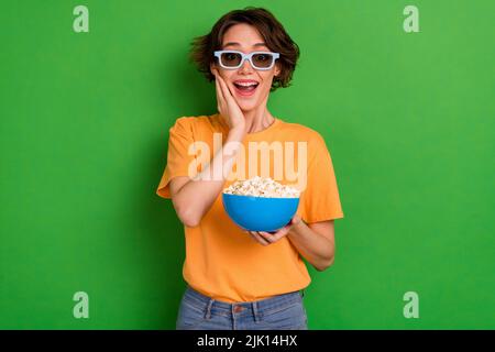 Photo of funky impressed young woman dressed orange t-shirt sunglass arm cheek eating pop corn isolated green color background Stock Photo