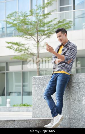 Cheerful Vietnamese man standing outdoors and checking messages in his phone Stock Photo