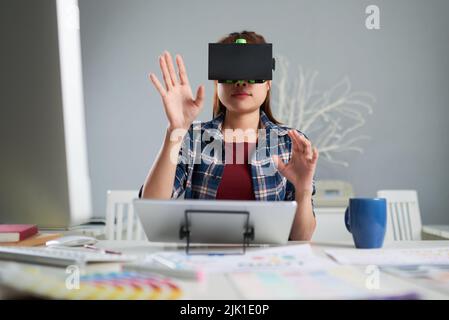 Young interior designer in casualwear using VR headset for work with 3d visualization while sitting at desk of spacious office Stock Photo