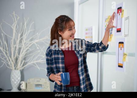 Cheerful Asian designer standing at whiteboard with cup of coffee in hand and brainstorming on start-up project, interior of stylish studio on background Stock Photo