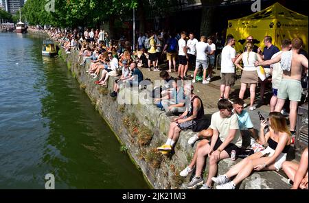 People enjoying the sun sitting along Bristol narrow quay wall on a sunny hot day during harbour festival, UK Stock Photo