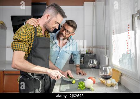Happy hipster gay couple in love hugging while standing in the kitchen. They are cooking dinner together at home and drinking wine. High quality photography. Stock Photo