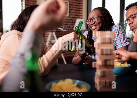 Multicultural happy friends enjoying drinking alcoholic beverage together while playing society games. Multiethnic joyful people sitting at home in living room while relaxing with leisure activity. Stock Photo