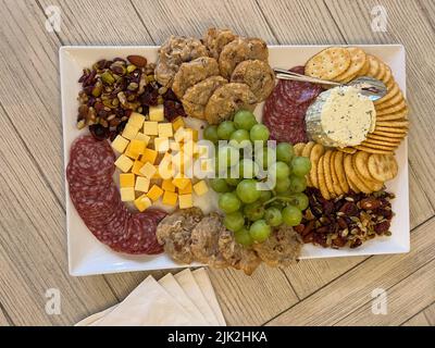 A charcuterie platter of cheese, grapes, nuts, sausage, cookies, crackers and cheese spread on a white platter sitting on a table. Stock Photo