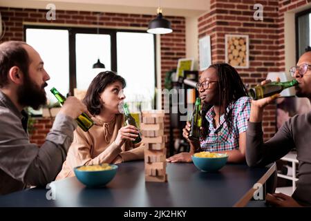 Multiethnic friends looking at each other while sipping beer and playing with wood blocks. Diverse people sitting together at home in living room while talking and enjoying fun leisure activity. Stock Photo