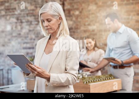 Mature female architect using a tablet to analyze designs in a modern office. Senior business woman with a blurred background of a young architectural Stock Photo